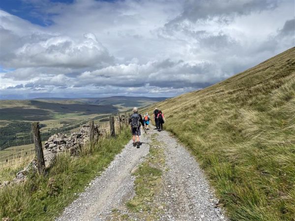 A group of ramblers walk along a gravel footpath along the Yorkshire countryside, on a cloudy day. They walk away from the camera towards the horizon. Clouds roll across the hills and green hills expand either side of the footpath